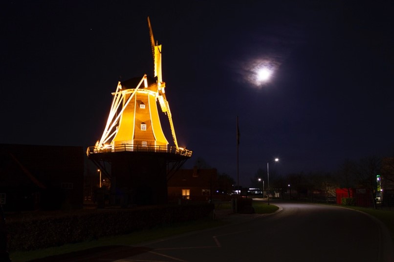 Verlichte molen in het donker langs een zwak verlichte weg. De maan schijnt. Zie bijschrift onder de afbeelding